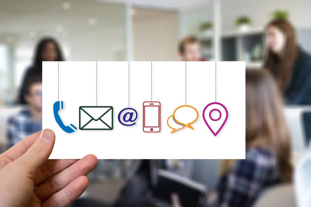 hand holding sign with icons including phone, envelope, @ symbol, cell phone, chat boxes, and map pin with blurry office in background to illustrate effective communication on web design project